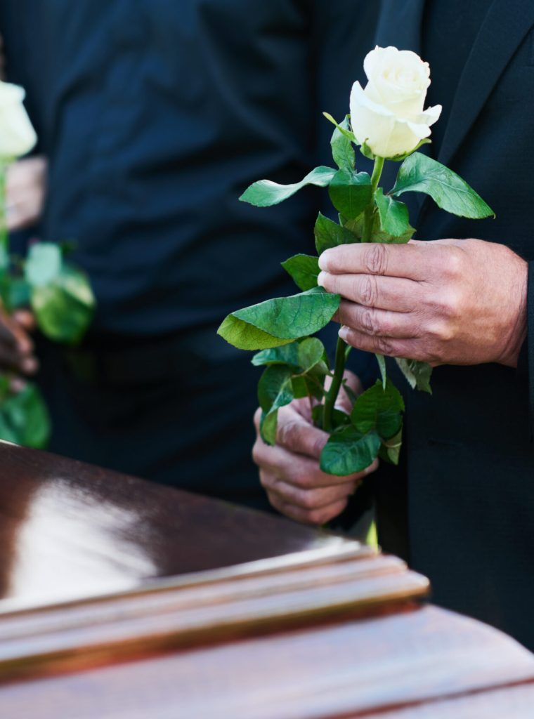 Hand of mature man in mourning clothes holding white rose while standing in front of coffin with closed lid at funeral of relative or friend